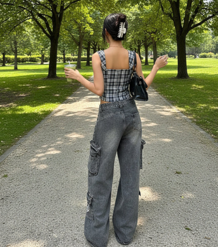 Person wearing a plaid top and gray pants, holding a drink and a black bag, standing outside.
